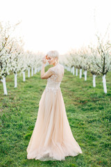 Fototapeta na wymiar Full-lengh portrait of young blond woman in long elegant dress in the spring apple garden, standing on green grass
