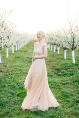 Fototapeta na wymiar Young beautiful blonde woman in blooming garden. Pretty bride in elegant peach dress, posing on the background of blooming trees and laghing