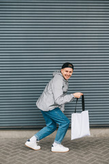 Funny young man with eco bag in his hand is having fun on the background of the gray wall, looking into the camera and smiling. Eco Friendly Positive guy crouches with a smile on his face