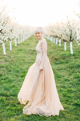 Young smiling woman in long elegant wedding dress walking outdoors on blossoming trees background. Cheerful bride walking outdoors in spring garden