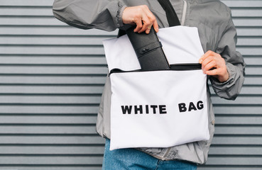 Young man stands outside in stylish clothes against a gray wall background and puts a wallet in a trendy white eco bag.Guy wears a reusable shopping bag, pulls out his wallet. Close photo. Background