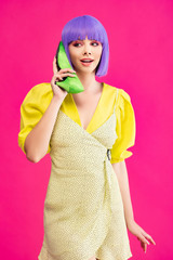 attractive pop art girl in purple wig holding green shoe as phone, isolated on pink