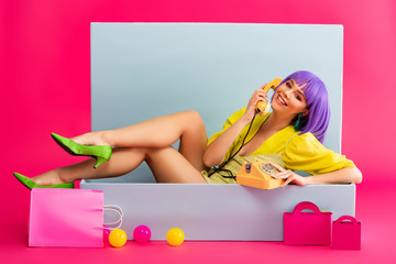 smiling girl in purple wig as doll talking on vintage phone while sitting in blue box with balls...