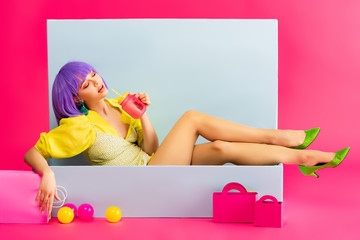 beautiful girl in purple wig as doll drinking from jar while sitting in blue box with balls and shopping bags, on pink