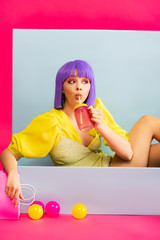 pop art girl in purple wig as doll drinking from jar while sitting in blue box with balls and shopping bag, on pink