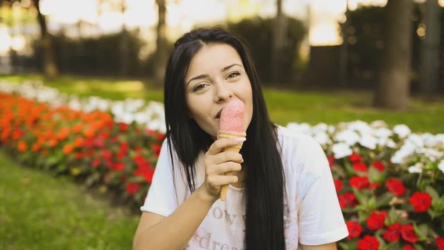 Young beauty woman is eating ice cream in HD VIDEO. She is smiling and licking pink gelato in front of flower bed in city park. Low depth of field and blurred background.