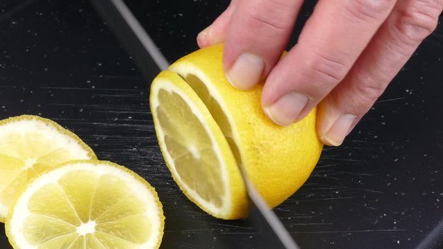 cutting lemon fruit into slices on a black cutting board with a kitchen knife closeup 4k