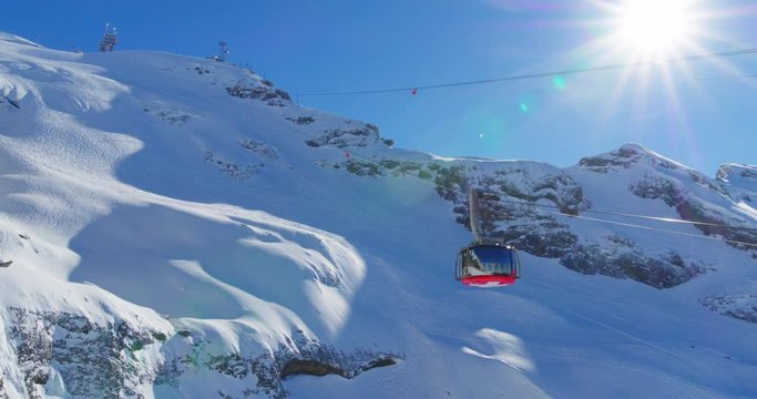 Ski lift slowly going up in the Swiss Alps. Sun is clearly visible above the snow covered mountaintops. Camera flare. Lens flare. Captured by drone.