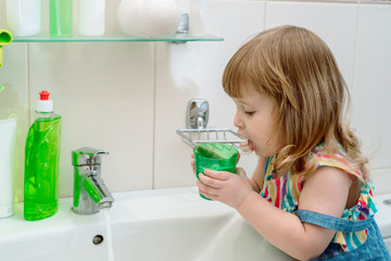 Morning hygiene in the bathroom. Baby the girl washes, brushes her teeth, rinses her mouth with water, and wipes herself with a towel. Smiling young girl brushing teeth and holding glass of water