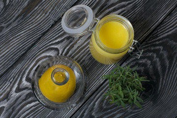 Lemon Kurd in a glass jar. Nearby is half a lemon in a container with a glass lid. A bunch of rosemary in a glass. Against the background of brushed boards.
