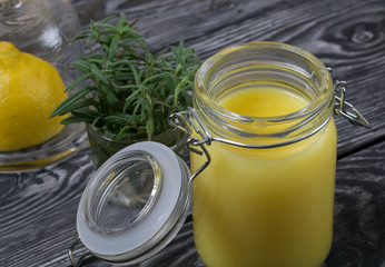 Lemon Kurd in a glass jar. Nearby is half a lemon in a container with a glass lid. A bunch of rosemary in a glass. Against the background of brushed boards.