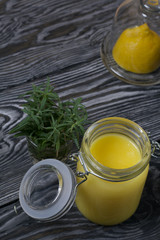 Lemon Kurd in a glass jar. Nearby is half a lemon in a container with a glass lid. A bunch of rosemary in a glass. Against the background of brushed boards.