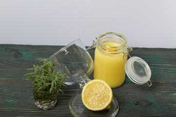 Lemon Kurd in a glass jar. Nearby is half a lemon in a glass container with a lid and a bunch of rosemary in a glass. Stand on brushed boards, on a white background.