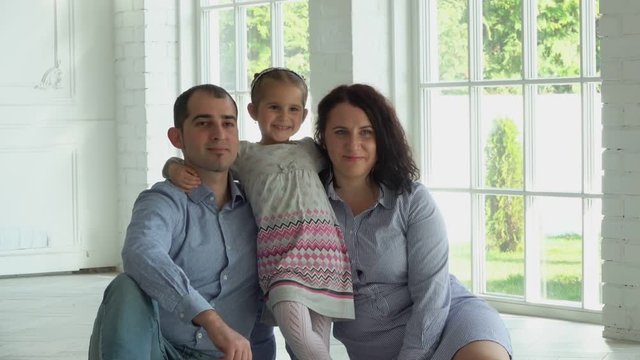 Photographer is taking pictures of a beautiful family with a cute smiling daughter in a sunlit wide-window white studio