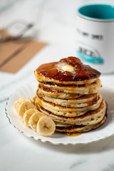 Hot american pancakes with bananas, melted butter and apricot jam on the white table. Mug, glasses and envelope background