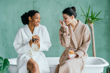 Cheerful pretty multiracial young ladies friends wear silk robes having fun hold glasses with champagne sit on bed in hotel, happy diverse girls bride and bridesmaid celebrate bachelorette spa party - obrazy, fototapety, plakaty