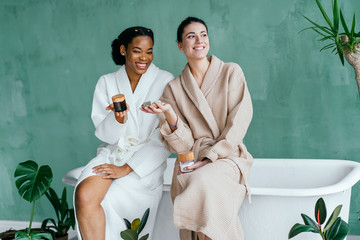 Portrait of two cheerful multiethnic women. Beautiful women in cozy bathroom, green background,many pots with plats, green leaves, flawless skin. Diverse friends.
