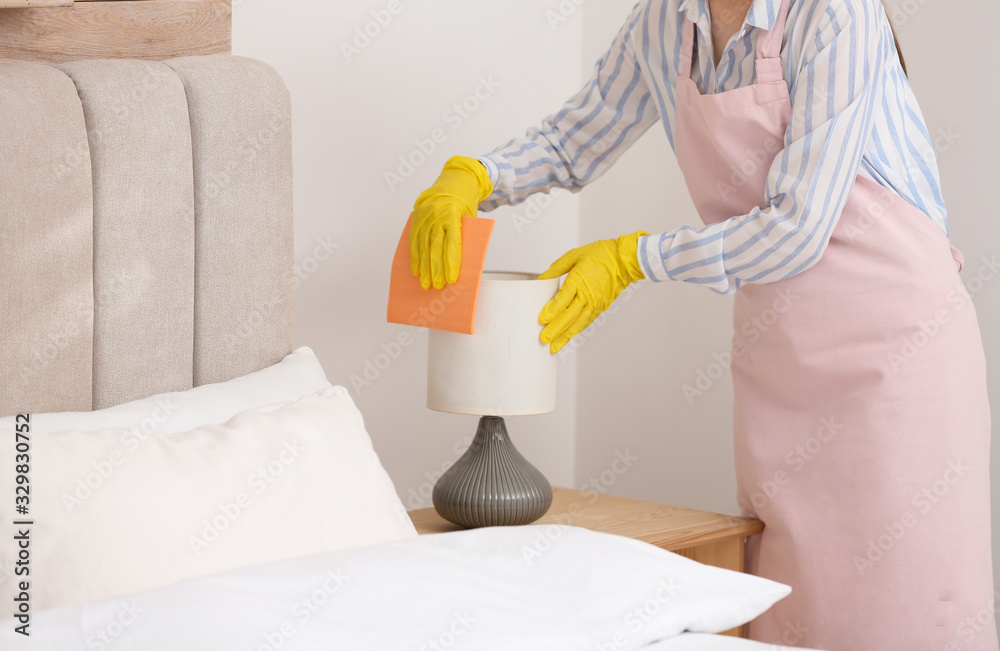 Sticker young chambermaid wiping dust from lamp in bedroom, closeup