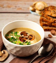 Hot tasty mushroom cream soup decorated with green onion and fried mushrooms in a white bowl on a brown kitchen towel with bread and fresh champignon background