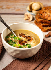 Tablespoon in a white bowl with hot tasty mushroom cream soup decorated with green onion and fried mushrooms on a brown kitchen towel with bread and fresh champignon background. Close up