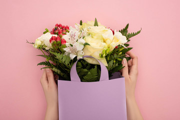 cropped view of woman holding bouquet of flowers in violet paper bag on pink background
