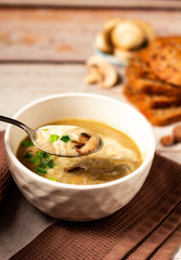Tablespoon with hot tasty mushroom cream soup decorated with green onion and fried mushrooms in a white bowl on a brown kitchen towel with bread and fresh champignon background. Close up