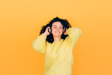 young beautiful girl with curly hair listens to music on headphones in a yellow sweater on a yellow background