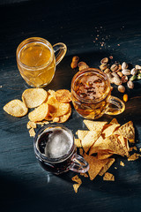 three glasses of expensive craft beer, classic and unfiltered and dark in a glass on the table with a snack of peanut and pistachio chips and nachos