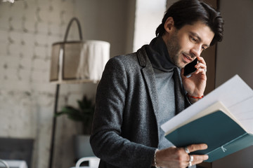 Image of young businessman holding papers and talking on cellphone in cafe
