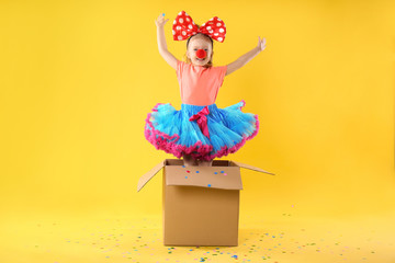 Little girl with large bow and clown nose jumping out of cardboard box on yellow background. April...