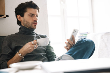 Image of handsome young man drinking coffee and using cellphone in cafe