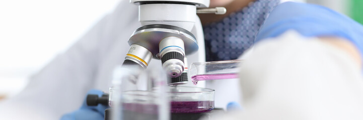 Close-up view of female biologist looking through microscope on purple liquid. Science research in...