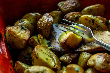 Fork cutting a baked potato wedge on the wooden spatula. Baked with spices, chopped garlic, olive oil and fresh chopped dill and green onion in a baking dish. Side view. Horizontal orientation