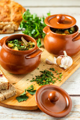 Clay pots with baked potatoes and chicken meat with pumpkin and choped greens on a wooden plank. With ca in the foreground. Garlic and bread on the table. Vertical orientation