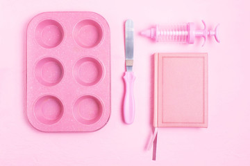 Cupcakes baking dish, a spatula, a recipe book, a towel on a pink concrete background. Monochrome. Copy space.