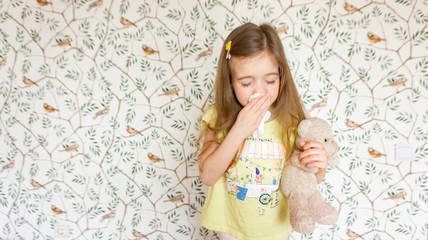 Obraz na płótnie Canvas A charming girl with a teddy bear gets sick and sneezes and coughs into a napkin. Symptoms of colds and flu. Allergy to pollen in a child. Allergic runny nose in a child.