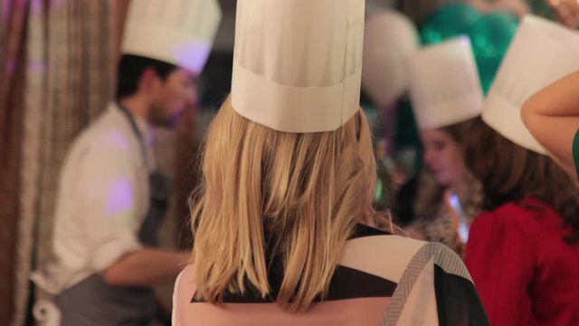 Master class of cooking. Blonde female party guest wearing chef hat in the middle. Anonymous shot with other people in blurry background