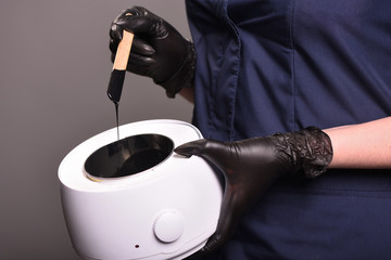 Black wax for depilation on a stick is held by a female hand in a black glove on a gray background