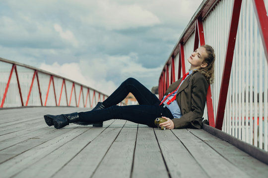 An attractive young man with long blonde hair, in berets and a military coat is leaning on a railing bridge, in his hand is a green flask.