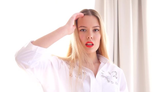 Portrait of a young white-haired girl by the window. Pretty young girl in a white shirt near the window. In the video, the girl poses against a large window. Light room.
