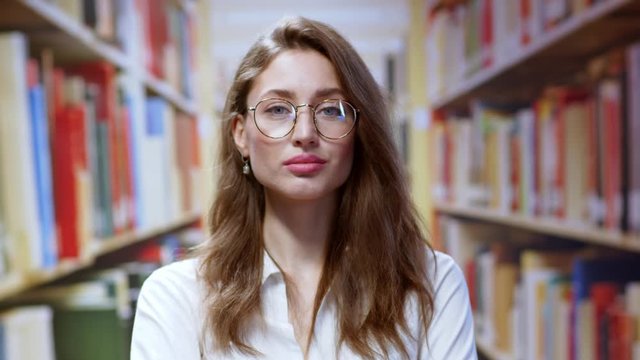 Young caucasian woman librarian wearing glasses looking smart and beautiful standing in row of bookshelves at modern library. Education, success concept.