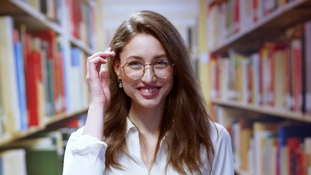 Real portrait of attractive smart caucasian young woman wearing glasses and braces smiling happy excited while staying at library bookshelves background.