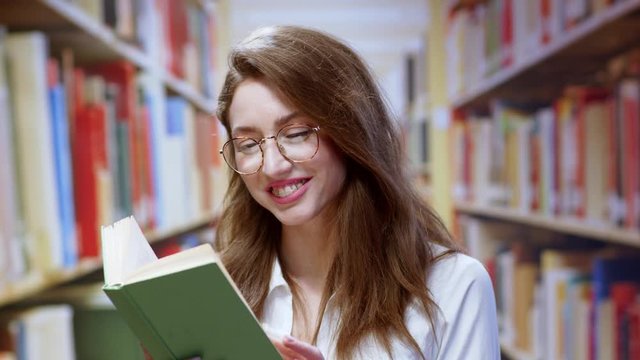 Pretty young woman librarian reading a book finds something good smiling cheerful satisfied enjoying her study at bookshelves library background.