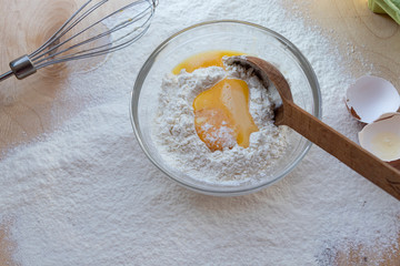 Cooking dough. A raw egg. Stir the protein with the yolk. Sifting flour through a sieve on a wooden background. Glass bowl with mix, towel and wooden spoon.