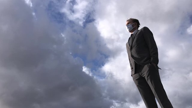Solitary businessman standing outdoors under the weather wearing a surgical face mask in isolation with time-lapse clouds