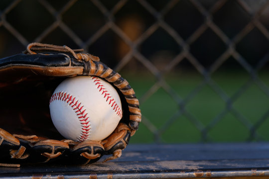 Baseball in glove close up with dugout chain link fence blurred background, copy space beside new ball.