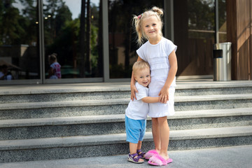 Two cute adorable caucasian blond little siblings standing and hugging on entrance residential building. Young sister and toddler brother enjoy playing together outdoors. Family love, care support