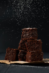 Stack of a brownie pieces on a grey table with dark background with sugar powder snow.