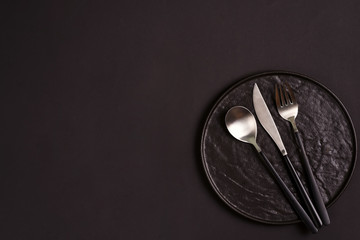 Black handmade ceramic plate served fork, spoon and knife on the same color background.