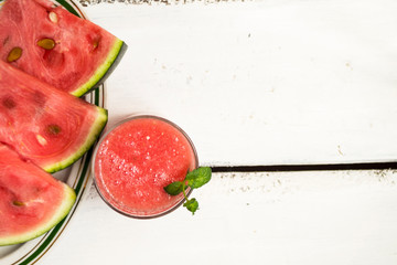 Slices of ripe watermelon on a plate and red watermelon juice in a glass on a wooden white background - top view. There is a place for text.